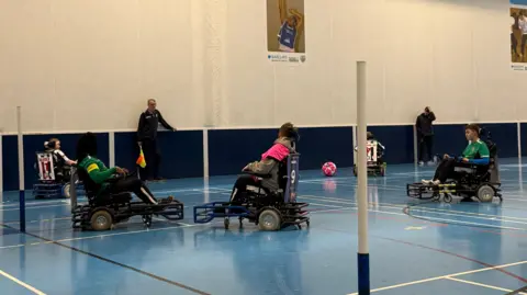 Supplied A few players sat in power chairs in a sports hall, they're focussed on the ball and an official with a flag watching on