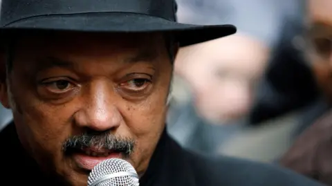 El reverendo Jesse Jackson, de Reuters, habla por un micrófono. Lleva sombrero y traje negros.