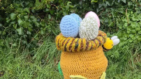 Carnon Downs Crafters A crocheted Easter-themed bollard cover. The yellow crocheted cover depicts an egg nest with four colourful eggs on top. A crocheted bee is stitched onto the side of the cover. Bushes are behind the bollard.