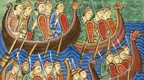 Getty A close-up of an illustration from a medieval manuscript showing Viking ships, with animal shaped prows, arriving in Britain. It shows men sitting side on in four boats with shields. The upper level shows the the whole boats, with their oars sticking into blue water; the lower level shows the men's heads and tops of their shields. Artist : Abbo of Fleury (c. 945-1004).