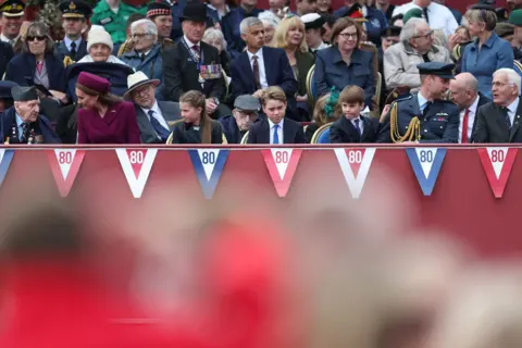 PA Media (L-R) Veteran Bernard Morgan, the Princess of Wales, Princess Charlotte, Prince George, Prince Louis and the Prince of Wales sit on the front row of benches of a crowd outside Buckingham Palace with bunting saying '80' at the front.