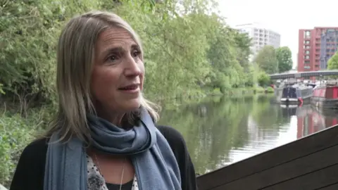 A woman stands in front of a canal with tower blocks in the background and greenery. She has blonde hair and has a light grey scarf.
