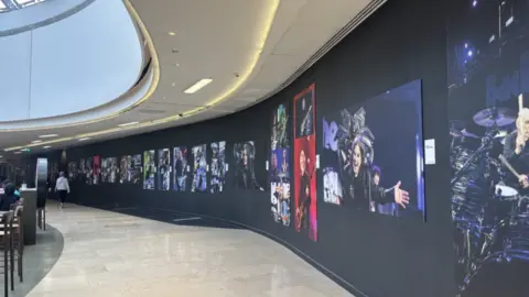 A photo of a photo exhibition of photos from the Back to the Beginning concert in Birmingham. It's on a black background and the floor is light. 