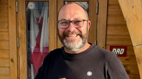 Marcus Baker is seen from his shoulders up. He is wearing a black T-Shirt and has a short beard. He is smiling at the camera.