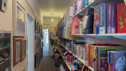 George King/BBC A view of the rescue centres hallway, which features long shelves filled with board games, toys, are other donated items. There are picture frames on the walls.
