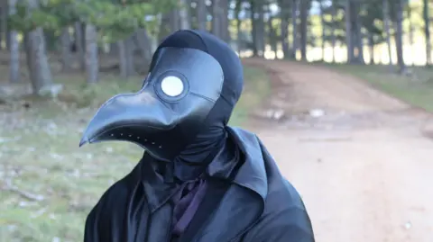 Generic image of person in black cloak and black mask with long beak in woodland