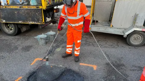 Simon Dedman/BBC A pothole repair in action. A man stands wearing bright orange safety clothing holding a long pipe and hose to heat the asphalt. There are vehicles behind him and two metal buckets.