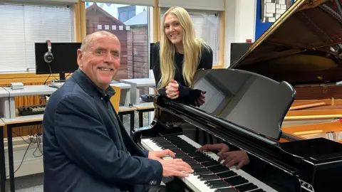 Adam Reece, a man in a dark blue jacket sitting at a black piano, looking at the camera, with a blonde woman Emily Fern leaning against piano also looking at the camera