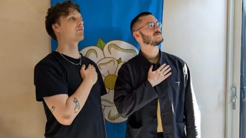 Two young men stand in front of a Yorkshire flag - a white rose on a blue background - hanging on the wall. Each has his hand on his chest, looking out of frame at the sky. It's an ironic pose reminiscent of football players lining up for their national anthem before a match.