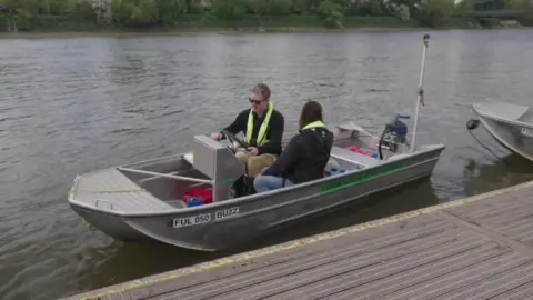 A high-angle shot of a man and a woman in a small, silver aluminium motorboat alongside a wooden pontoon. The man, wearing sunglasses and a high-visibility life vest, sits at the steering console while the woman sits facing forward near the back.