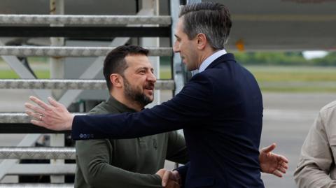 Simon Harris wearing a navy suit and Volodymyr Zelensky wearing a green jumper hug as they meet at Shannon Airport