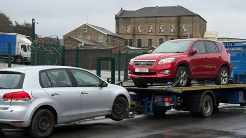 Bradford Council A truck with a red vehicle on top of it and a grey car being towed by it in Bradford