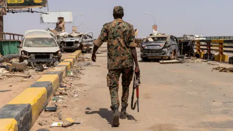 A lone soldier walks across a bridge. Bombed-out cars and rubble can be seen on the bridge.
