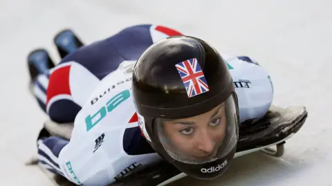 Getty Images Shelley Rudman on a sled racing down a track in her Skeleton race. She is wearing a white, blue and red Team GB outfit with a helmet. 