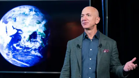 Jeff Bezos wears a grey suit an stands on a stage with his left hand out as he talks, with a graphic of the earth on a screen behind him 