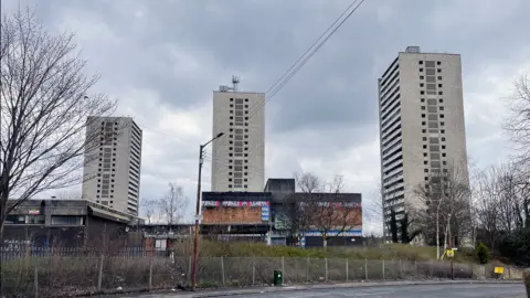 BBC Three high rise blocks of flats behind a fence. There are dilapidated buildings in the foreground