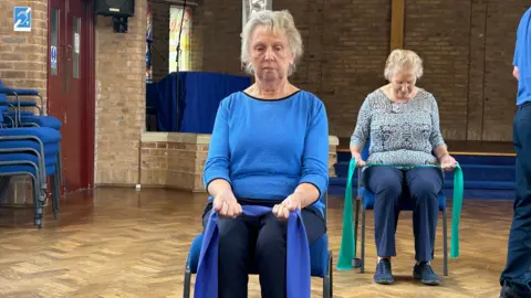 A woman wearing a blue jumper sitting on a chair holding a blue exercise band