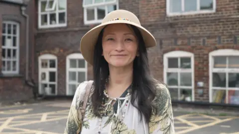 A woman wearing a wide-brimmed hat and patterned top standing in front of a brick building with large windows.