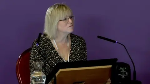 Southport Inquiry Joanne Hodson, who has shoulder length blonde hair, a fringe and glasses, sits at a witness table in front of a screen and a black microphone in front of a purple screen. 