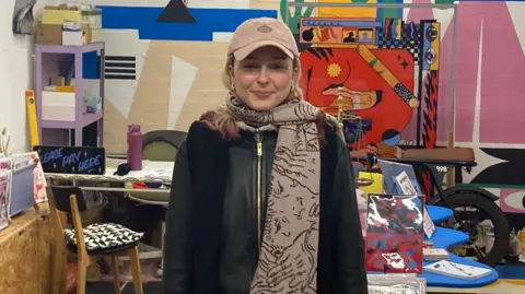 Daisy is a young woman with blonde hair and a beige baseball cap. She is wearing a matching scarf and a black leather jacket. She is centre of frame, smiling and the room behind her is full of colourful arts and crafts work.