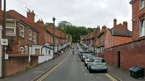 View of Vine Street in Lincoln. It is a row of red brick terrace houses along a hill with trees at the end. Cars are parked alongside the right-hand side of the road. 