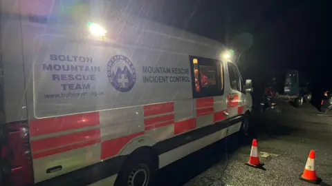 Bolton Mountain Rescue A mountain rescue vehicle parked by some traffic cones in the dark at a car park. Rain can be seen lashing against the side of the vehicle. 