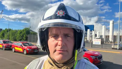 A firefighter wearing a white helmet stands in the foreground, facing the camera, while smoke rises from an industrial site behind him. Red emergency vehicles line the road, and the scene appears to be an active emergency response under a bright, cloudy sky.