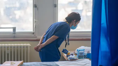 A nurse wearing blue scrubs looks down at a wooden table next to a patient's bed. The bed is empty and has been made up. A blue curtain hangs next to the bed.