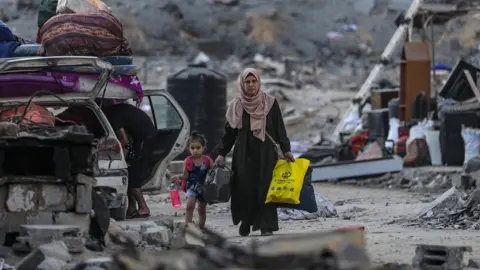 EPA/Shutterstock A Palestinian woman and child walk through the rubble of Gaza City holding hands after being internally displaced by an Israeli airstrike. The woman wears a headscarf and carries plastic shopping bags. The young girl beside her carries a pink water bottle. Debris lines the street.