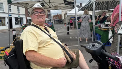 Gene Hicks sits on his mobility scooter and looks to the right towards the camera. He is wearing a yellow polo shirt and a pork pie hat with the work 'Lanzarote' on it. He has a satchel slung over one of his shoulders. The street market is visible with traders and shoppers in the background