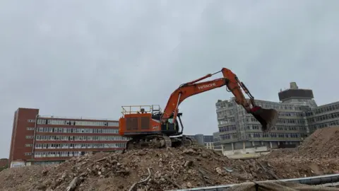An orange demolition vehicle sits on top of a pile of rubble with two large office buildings in the distance o a grey day.