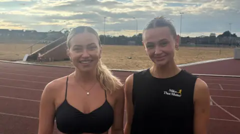 Two women standing on a running track, smiling at the camera. The one on the left has long, blonde hair tied back and is wearing a black sports bra. The one on the right has short, brown hair tied back in a bun. She is wearing a black vest, with a white and orange Miles That Matter logo on the chest.