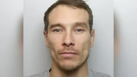 A close-up police mug shot of Lewis Moore on a white background. He has brown hair and a stubbly beard, and is wearing a grey T-shirt. 