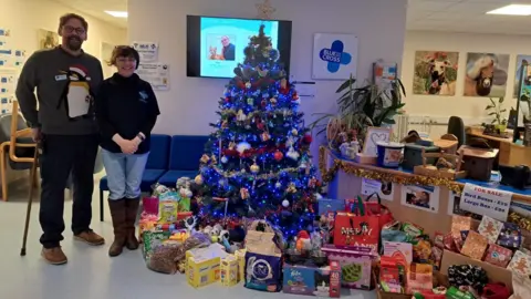 Clare Phillips/BBC Charles White and a Blue Cross staff member smile at the camera. To the right of them is a Christmas tree surrounded by presents that have been donated for the animals at the centre.