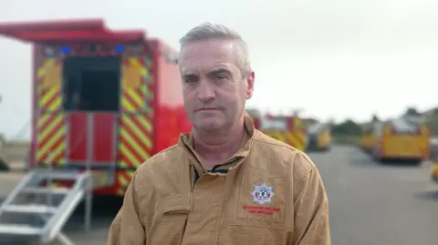 Peter Blaney, a man with grey hair and wearing a brown NIFRS uniform. There is a red and yellow fire engine in the background.