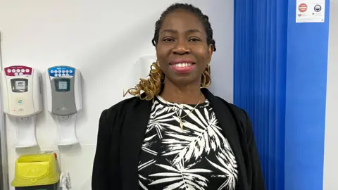 Bola Owolabi smiling at the camera wearing a black and white left patterned dress and a black jacket.