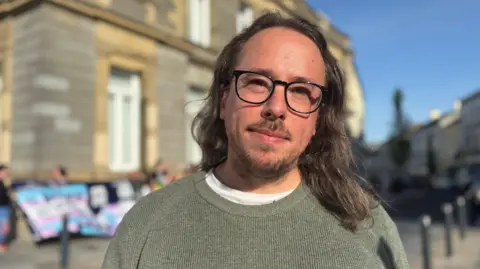 A long-haired man wearing glasses. He is wearing a crew neck jumper and behind him you can make out a small protest and a trans flag.