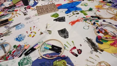 A white tablecloth covered with arts and craft supplies - pens, paints, wool, letter blocks - and artwork. 