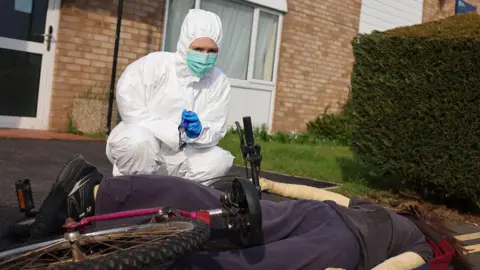 A woman in a white forensics suit and blue mask crouches outside a house next to a women's dummy lying facedown on the pavement. A bike is on top of her.