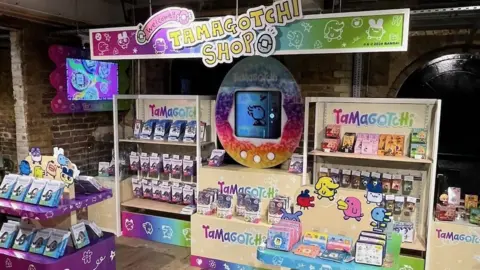 Tamagotchi शॉपमध्ये मागील बाजूस एक भव्य तमागोचीसह एक डिस्प्ले आणि अनेक शेल्फ् 'चे अवशेषांवर बरीच उपकरणे आहेत