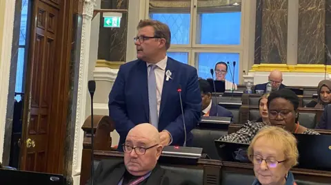 Councillors sit in tiered benches with microphones in front of them during a budget meeting in a council chamber. One man is on his feet. He is wearing a navy suit, pale blue tie, white shirt and dark-framed glasses. He has short brown hair and his face is in profile. Other members look down at laptops and other devices.