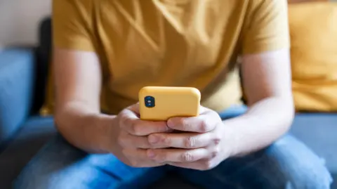 A close up of a person holding a yellow phone, sitting on a blue sofa, wearing jeans and a yellow t-shirt.
