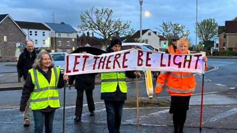 Three people wearing fluorescent vests are holding a banner saying ''Let there be light''