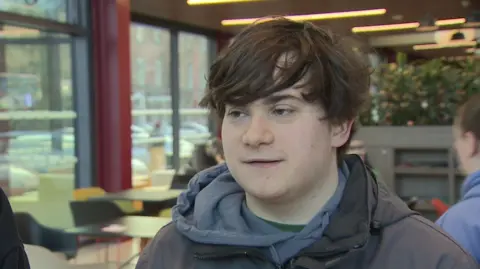 A young man in a grey hoody. He has longish brown hair swept over to the side.