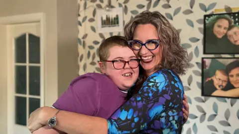 A woman and a boy are hugging. The woman is wearing a blue and purple patterned blouse and the boy is wearing a pink t-shirt. On the wall behind them, there are photos of the woman and the boy when they were younger.