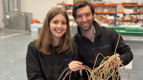 Olivia Simpson and Dr Mattia Parati are pictured smiling holding spaghetti-like strands stood next to each other. Ms Simpson has shoulder length brown hair while Mr Parati has dark brown hair slicked to the side. Both are wearing a black jacket.