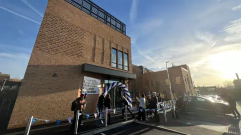 The building is sand coloured and modern looking. There's a balloon arch over the door in the NHS colours of white and blue. There is also blue and white bunting up over the entrance. 