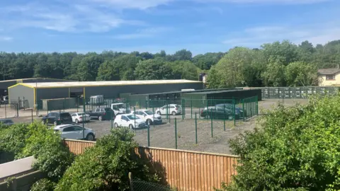 Photograph of the back of the Europa Trading estate in Bolton. The image shows fencing, shopping containers and a car park which has been built on the land.
