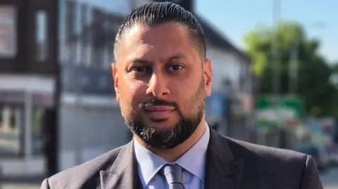 Shaz Saleem facing the camera. He has black hair, slicked back and a black beard and moustache. He is wearing a grey suit with a blue shirt and tie