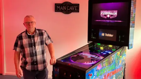 A pink-lit room, with a man in a chequered shirt stood next to a large multi-coloured arcade machine, with vertical and horizontal screens.